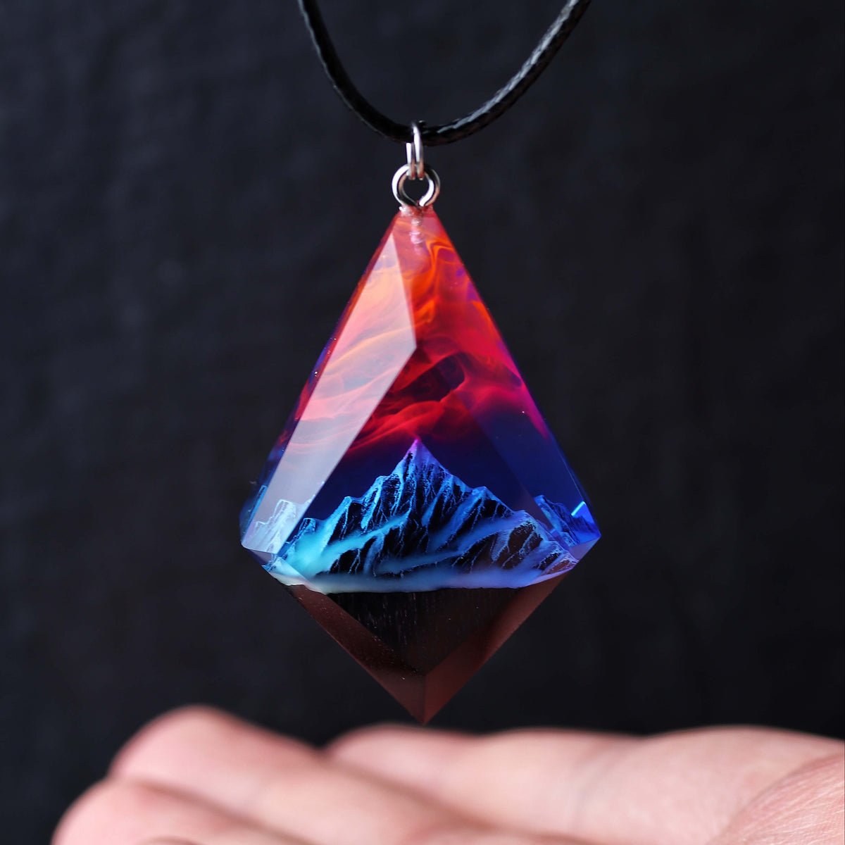 Wood resin necklace with fire and nebula sky over blue snowy mountains, faceted rhombus pendant held in hand, handmade jewelry