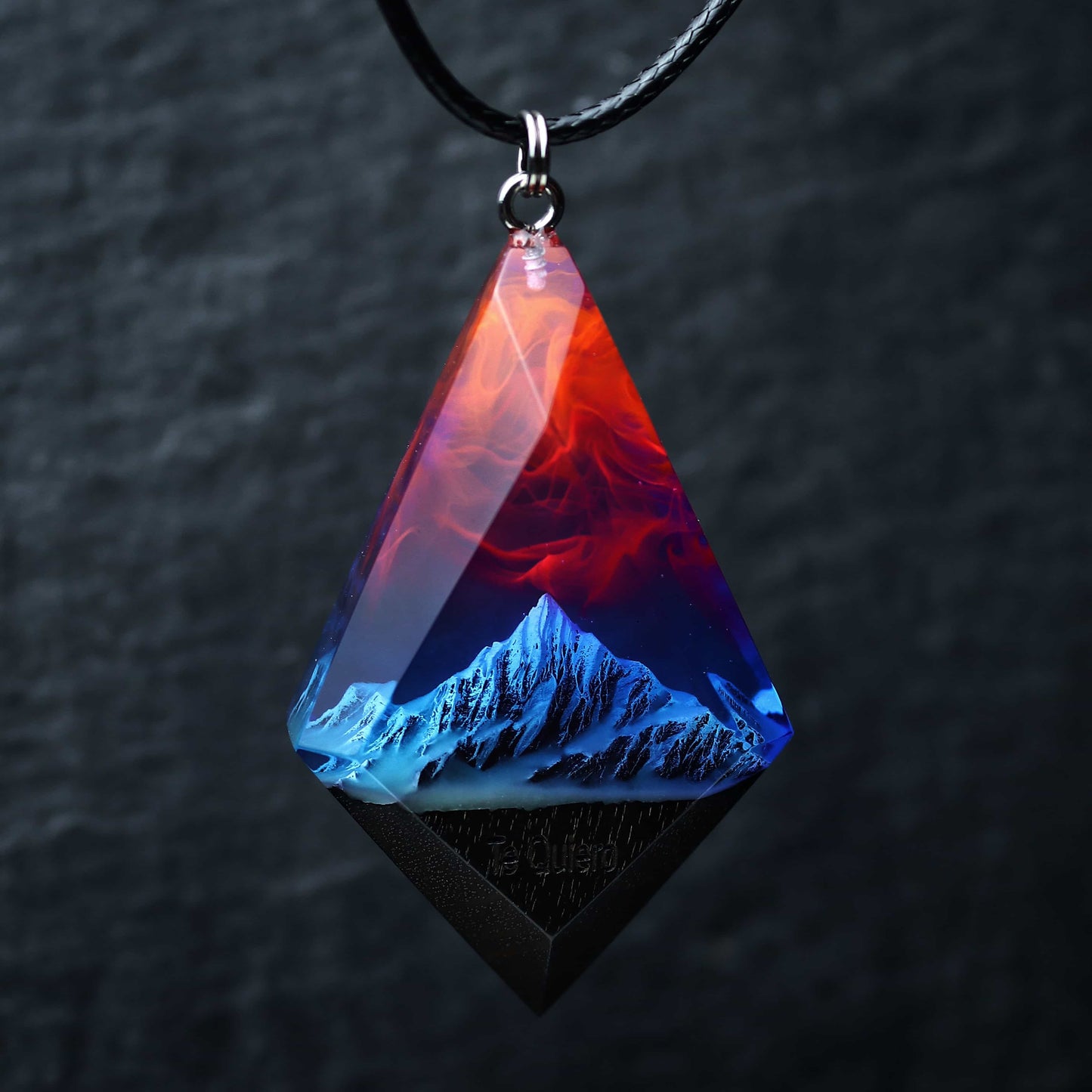 Wood resin necklace with fire and nebula sky over blue snowy mountains, faceted rhombus pendant held in hand, handmade jewelry