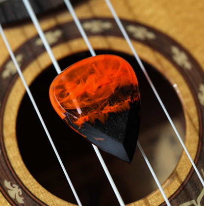 Wood resin guitar pick with orange fire design inside, handmade plectrum lying on guitar strings over the sound hole